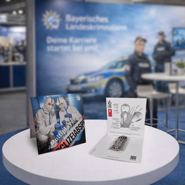 Bedruckte Pocketcard mit Motiv des Bayerischen Landeskriminalamts und Produktdarstellung aufgeklappt auf einem weißen Messetisch.