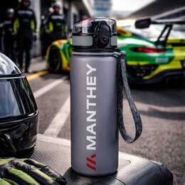 Graue Trinkflasche mit Manthey Logo steht auf einer Rennstrecke vor einem Sportwagen, Helm und Handschuhe liegen daneben